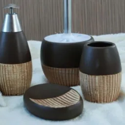 Zestaw łazienkowy Vulcano w odcieniach brązu: dozownik do mydła z metalową pompką, kubek na szczoteczki, mydelniczka z żłobieniami i pojemnik na szczotkę WC, ustawione na puszystym białym ręczniku.