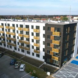 FACHMAN - Nowoczesny budynek wielorodzinny z białymi i grafitowymi elewacjami, ażurowe balkony w kolorze złota, widok z góry na parking i otoczenie.