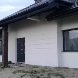 Nowoczesny dom jednorodzinny z białą elewacją i ciemną stolarką okienną, fragment wykończonej ściany z poziomymi dekoracyjnymi pasami, ceglana kolumna przy oknie, okno zabezpieczone folią ochronną.