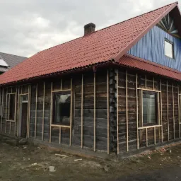 Dom z bali w trakcie przygotowywania do prac elewacyjnych: montaż drewnianej konstrukcji pod przyszłe ocieplenie, czerwony dach z blachy trapezowej, niebieski szczyt z dekoracyjnym wzorem.