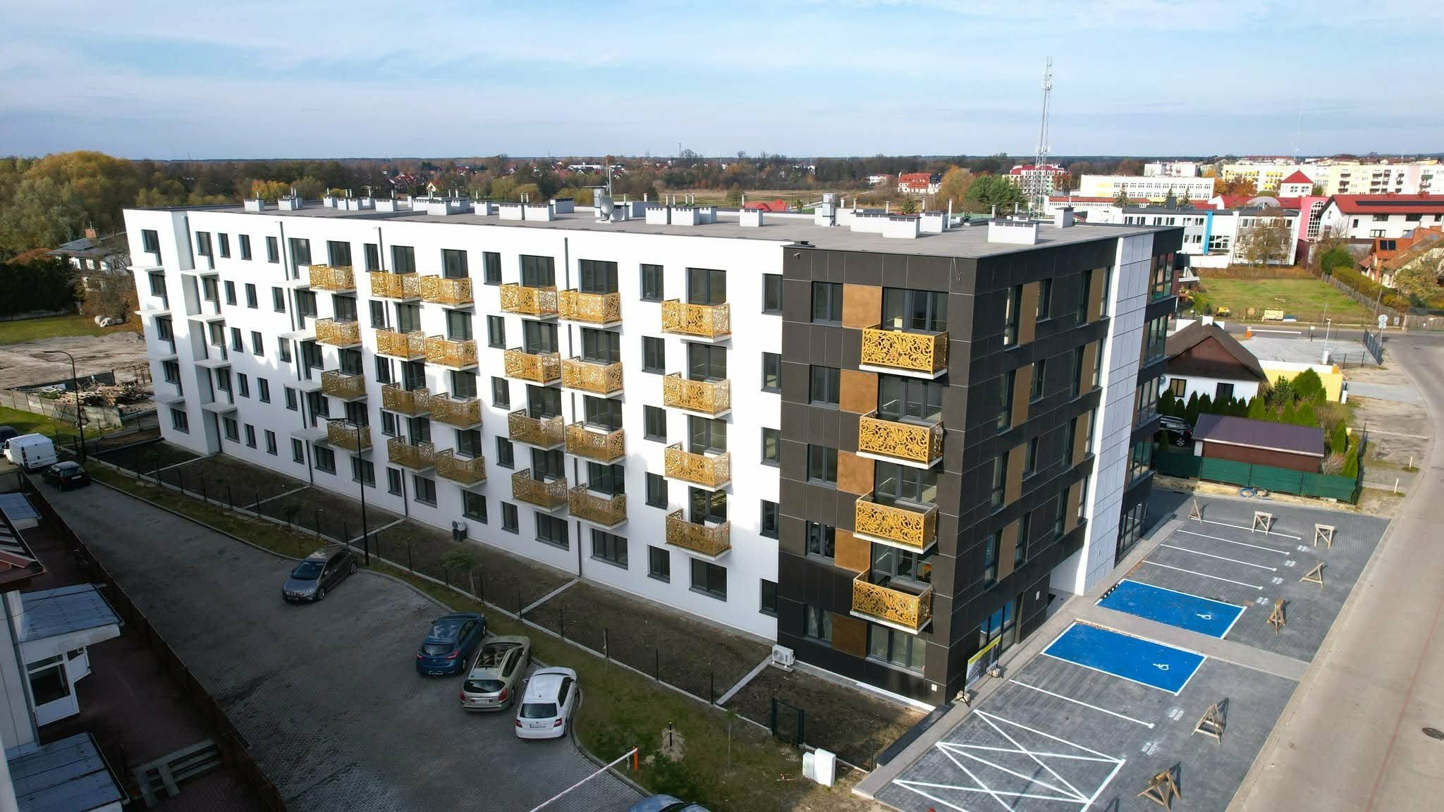 Nowoczesny budynek wielorodzinny z białymi i grafitowymi elewacjami, ażurowe balkony w kolorze złota, widok z góry na parking i otoczenie.
