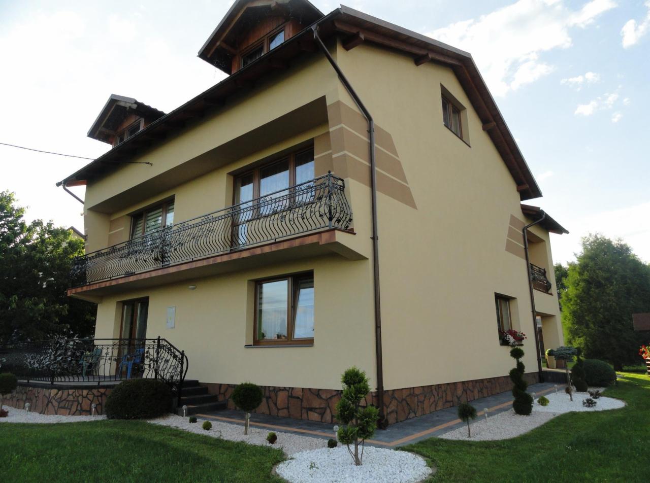 Dwupiętrowy dom z beżową elewacją, ozdobnymi balkonami z kutymi balustradami i elementami dekoracyjnymi w postaci poziomych pasów w odcieniach brązu, otoczony zadbanym ogrodem z białymi kamykami...