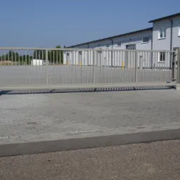 Szara, metalowa brama przesuwna z pionowymi szczeblami, zamykająca wjazd na posesję. Obok fragment ogrodzenia panelowego i budynek w tle.