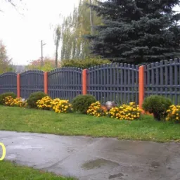 Grafitowe ogrodzenie sztachetowe z pomarańczowymi słupkami, obsadzone żółtymi kwiatami i zielonymi krzewami. Widok z chodnika w pochmurny dzień.