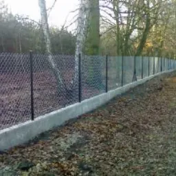 Długa siatka ogrodzeniowa na betonowej podmurówce, otoczona drzewami i liśćmi. Perspektywa z dołu ukazuje solidność konstrukcji i jej wkomponowanie w naturalne otoczenie.