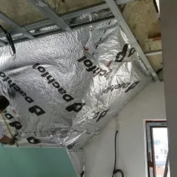 Montaż izolacji termicznej dachu z folii aluminiowej, widoczne profile metalowe i pracownik z miarką. Izolacja z nadrukiem, okno w tle.