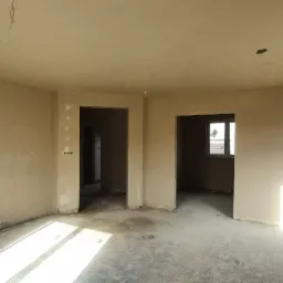 Wnętrze w stanie surowym, ściany pokryte świeżym tynkiem, widoczne otwory pod gniazdka elektryczne i wystające przewody z sufitu, posadzka betonowa przed wylewką, dwa przejścia do innych pomieszczeń.