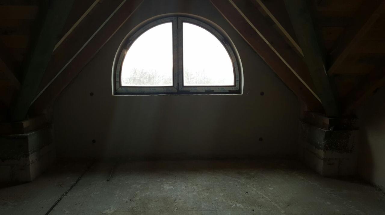 Poddasze ze świeżo otynkowanymi ścianami, widoczne drewniane belki stropowe i okno w kształcie półkola. Ściany szare, surowy stan wykończenia podłogi.