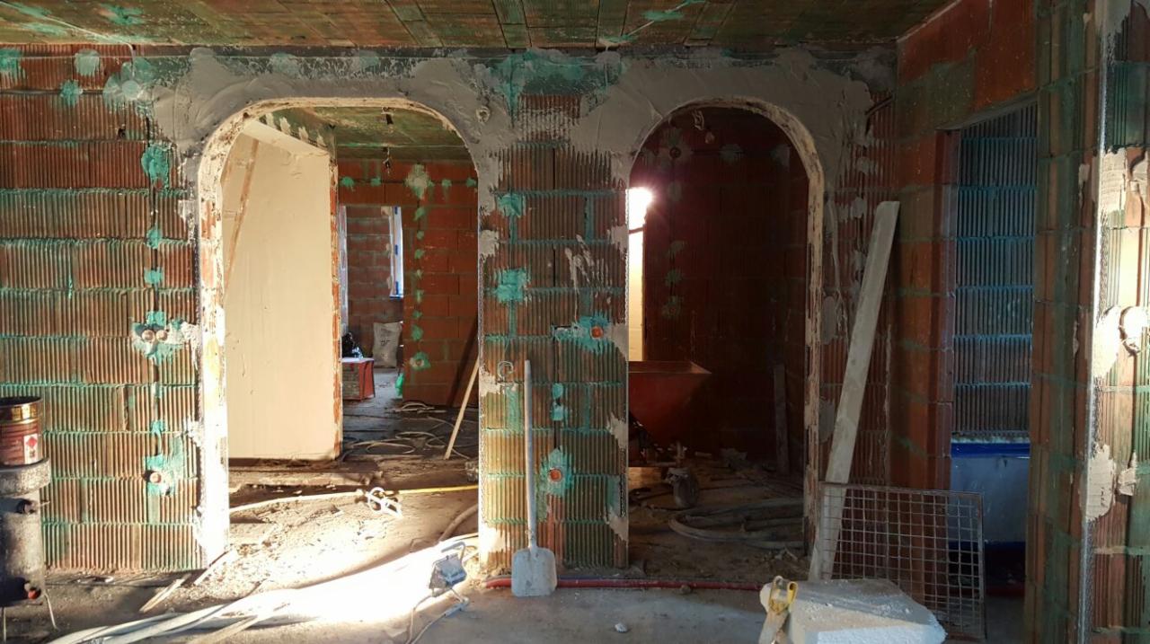 Wnętrze w trakcie tynkowania: widoczne ceglane ściany z zaznaczonymi miejscami na instalacje, łuki drzwiowe w trakcie obróbki tynkarskiej, narzędzia budowlane na podłodze.