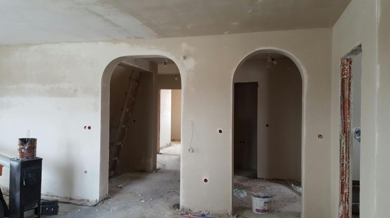 Wnętrze po tynkowaniu maszynowym: gładkie ściany z łukowatymi przejściami, widoczne ślady prac budowlanych i drabina oparta o ścianę. Nowe gniazdka elektryczne.