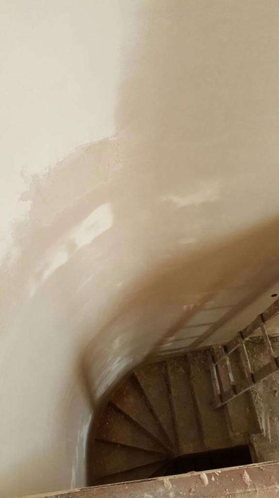 Świeżo nałożony tynk na zakrzywionej ścianie klatki schodowej, widok z góry na schody i rusztowanie. Gładka, jasna powierzchnia tynku w kontraście z surowymi schodami.