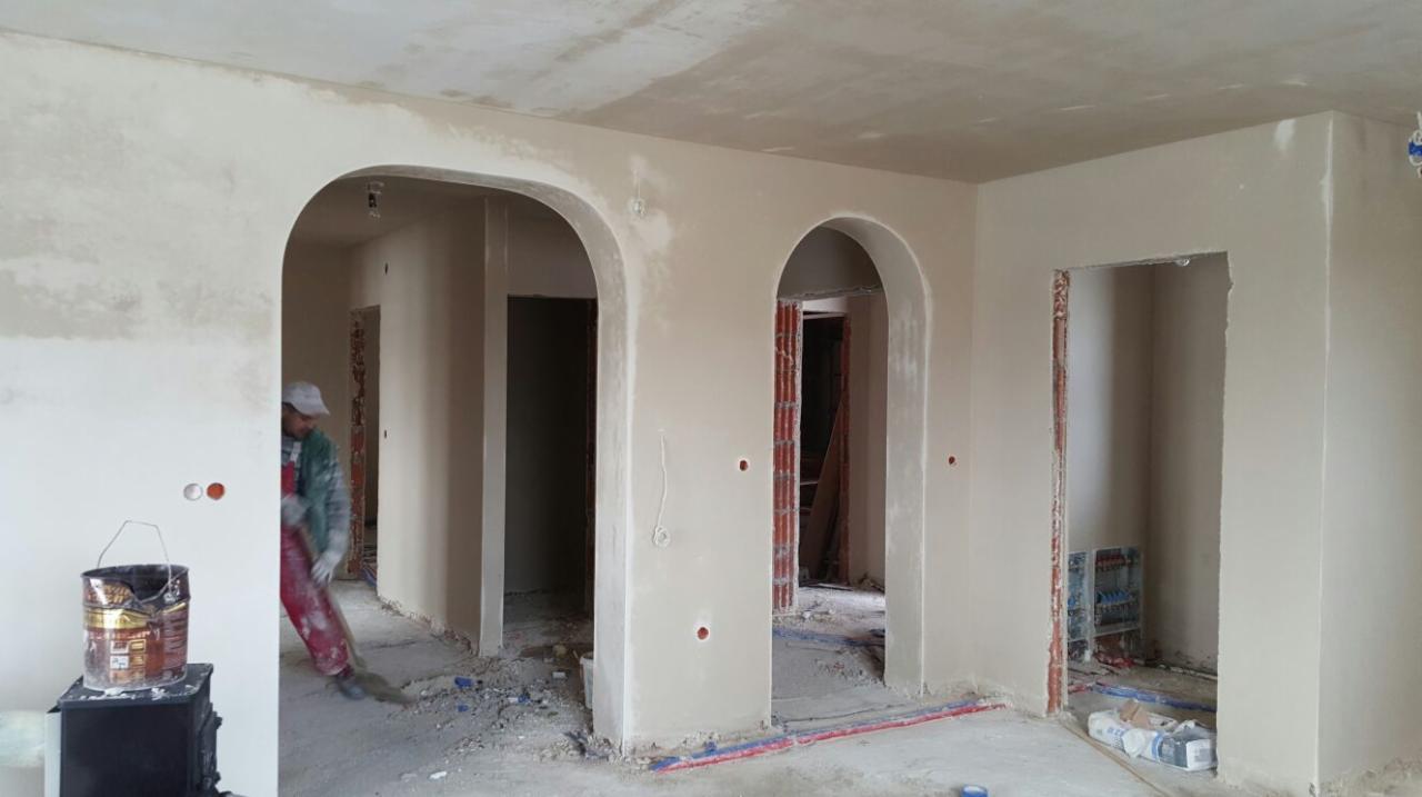 Wnętrze po tynkowaniu maszynowym, widoczne łukowe przejścia, instalacje w podłodze i pracownik w tle. Ściany gładkie, przygotowane do dalszych prac wykończeniowych.