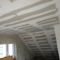Poddasze w trakcie zabudowy płytami gipsowo-kartonowymi, widoczne łączenia wzmocnione taśmą i pokryte szpachlą. Okno w tle. Skos dachu.