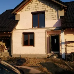 Dom w trakcie ocieplania styropianem, z widocznymi brązowymi oknami z roletami. Częściowo ocieplona elewacja, widoczne rusztowania i materiały budowlane.