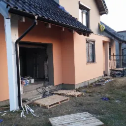 Dom jednorodzinny w trakcie prac elewacyjnych: świeżo otynkowana ściana w odcieniach beżu, widoczne rusztowania i materiały budowlane.