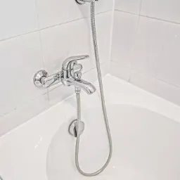 Bateria wannowa chromowana, zamontowana na białej ścianie z płytek. Wąż prysznicowy zwisa do wanny. Ujęcie z góry, widok na armaturę łazienkową po remoncie.