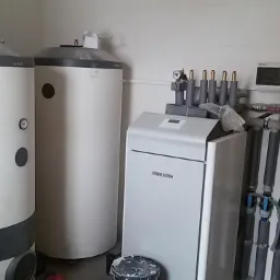 Dwa białe zbiorniki akumulacyjne z szarymi elementami i manometrami, obok nich skrzynka sterująca z rurami, instalacja w pomieszczeniu z białymi płytkami.