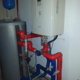 Instalacja z pompą ciepła Panasonic Aquarea 9kW, widoczne rury w kolorze czerwonym i niebieskim, zbiornik akumulacyjny i elementy sterujące.