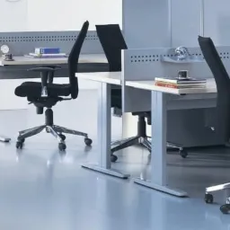 Nowoczesne biuro z jasnymi biurkami, czarnymi krzesłami obrotowymi i szarymi ściankami działowymi. Na biurkach lampki i segregatory. Minimalistyczny design.