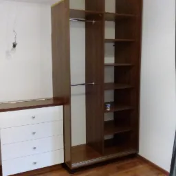 Montaż brązowej szafy wnękowej z białymi szufladami w pokoju z drewnianą podłogą i szarą zasłoną, widoczna żarówka bez klosza.