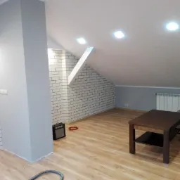 Wykończone poddasze z białymi ceglanymi ścianami, drewnianą podłogą, szarą ścianą, grzejnikiem i brązowym stolikiem kawowym, oświetlone punktowymi lampami sufitowymi.