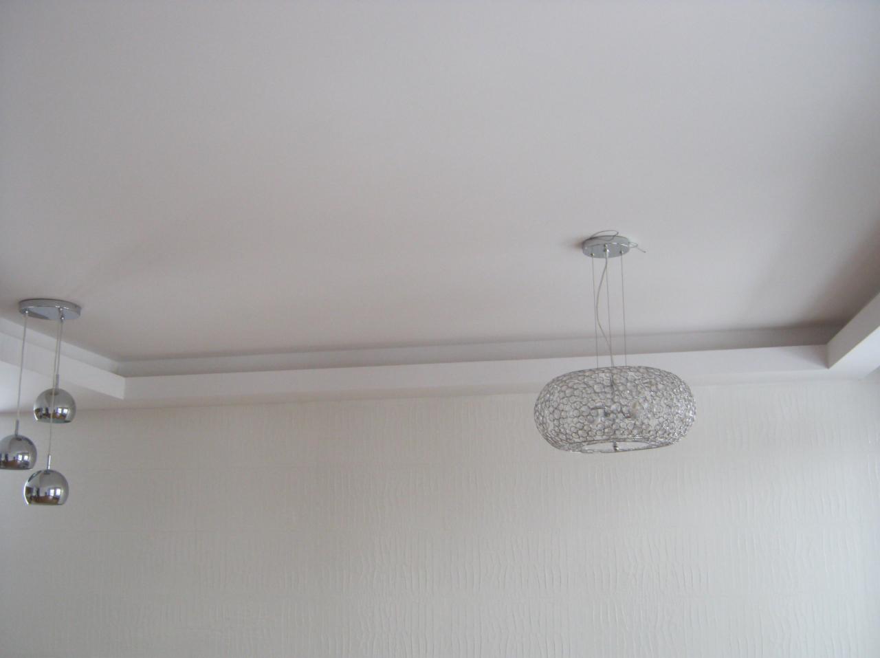 Jasne wnętrze z dwiema lampami sufitowymi: chromowana lampa wisząca z trzema kulami po lewej i okrągła, ażurowa lampa po prawej, biały sufit z dekoracyjnym obramowaniem i ściana z delikatną teksturą.