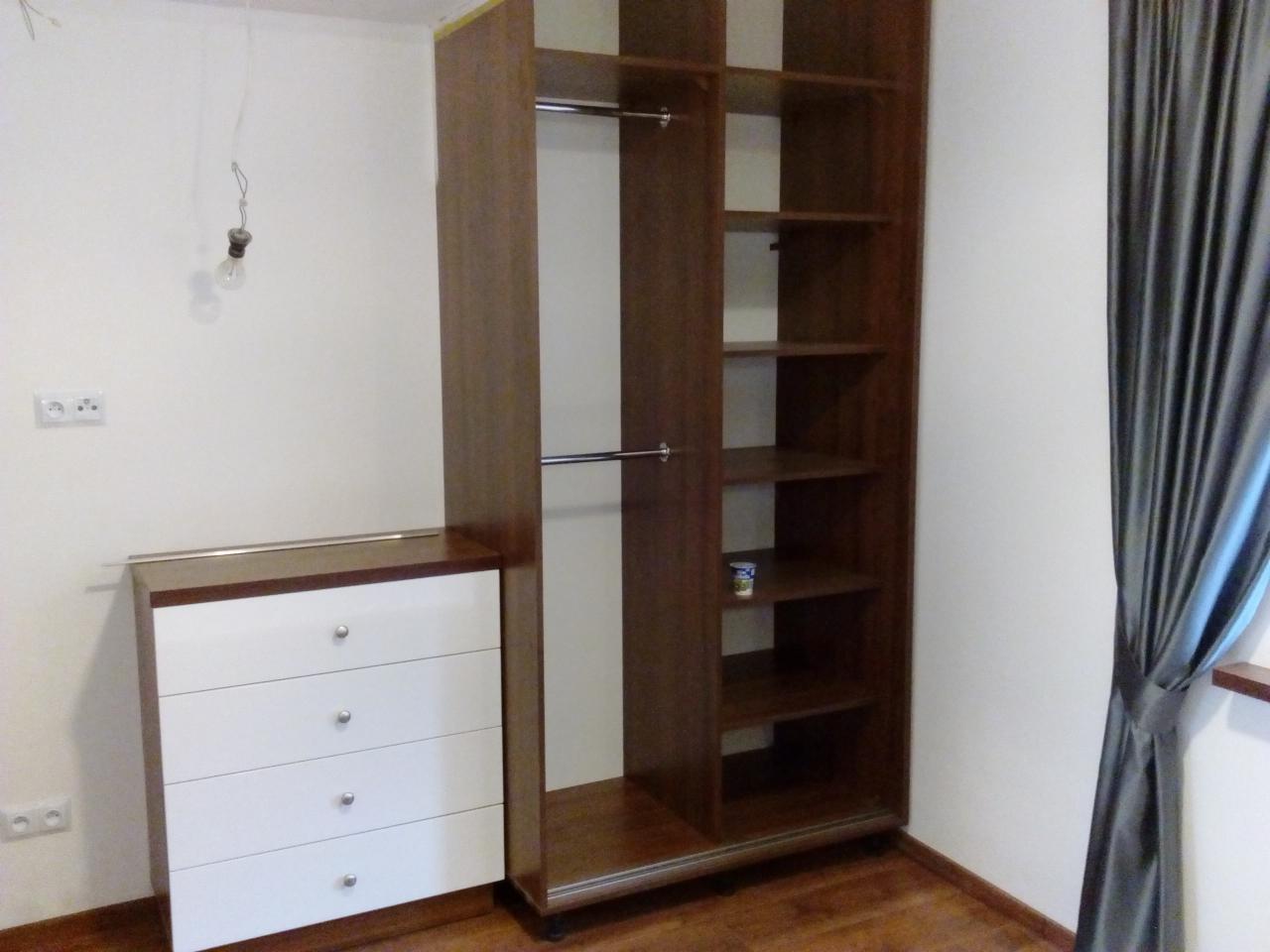 Montaż brązowej szafy wnękowej z białymi szufladami w pokoju z drewnianą podłogą i szarą zasłoną, widoczna żarówka bez klosza.