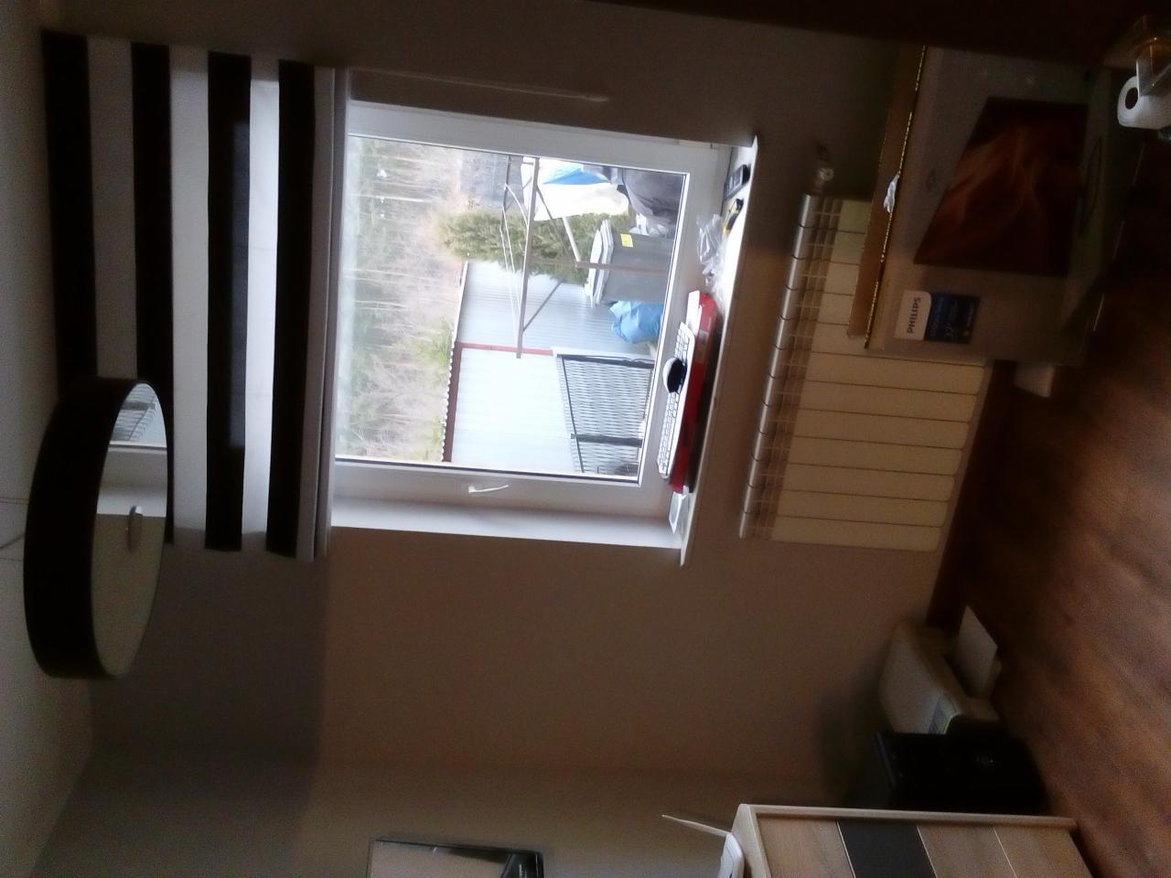 Pokój po remoncie: okno z roletami w czarno-białe pasy, grzejnik pod oknem, widok na zewnątrz z budynkiem gospodarczym w tle.