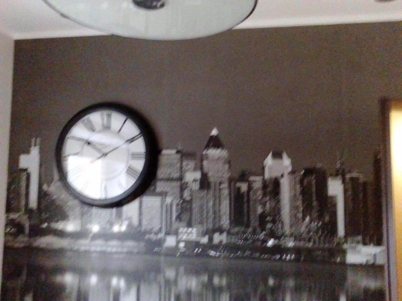Tapeta z motywem panoramy miasta w czerni i bieli, z okrągłym zegarem na ścianie w mieszkaniu w Warszawie. Widoczna lampa sufitowa.