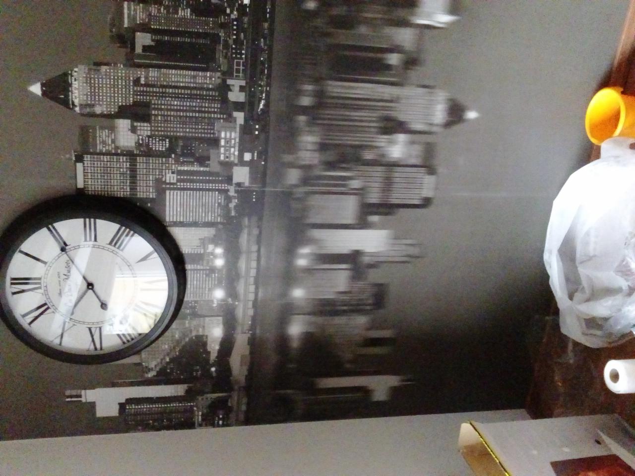 Ściana z czarno-białą fototapetą przedstawiającą panoramę miasta, obok wisi zegar, widoczny fragment biurka z kubkiem i reklamówką.