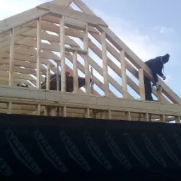 Montaż drewnianej konstrukcji dachu domu w trakcie budowy, widoczne krokwie i słupy, dw&oacute;ch pracownik&oacute;w w odzieży roboczej, czarna membrana z napisem MASTERMAX na ścianie, błękitne niebo w tle.