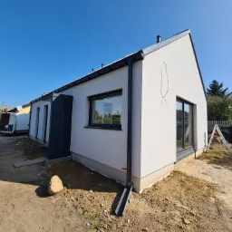 Nowoczesny dom parterowy z białą elewacją i czarnymi ramami okiennymi, widoczne ślady po pracach elewacyjnych, teren wokół domu w trakcie wyrównywania, drabina oparta o ścianę, błękitne niebo.