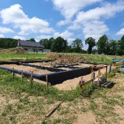 Przygotowanie do wylewki fundamentów pod dom jednorodzinny, widoczne szalunki i folia izolacyjna, w tle częściowo ukończony budynek oraz biały samochód dostawczy.