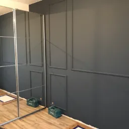 Ciemnoszara ściana z dekoracyjnymi panelami ściennymi, widoczne narzędzia i materiały przygotowane do dalszych prac wykończeniowych, odbicie w lustrzanych drzwiach szafy.