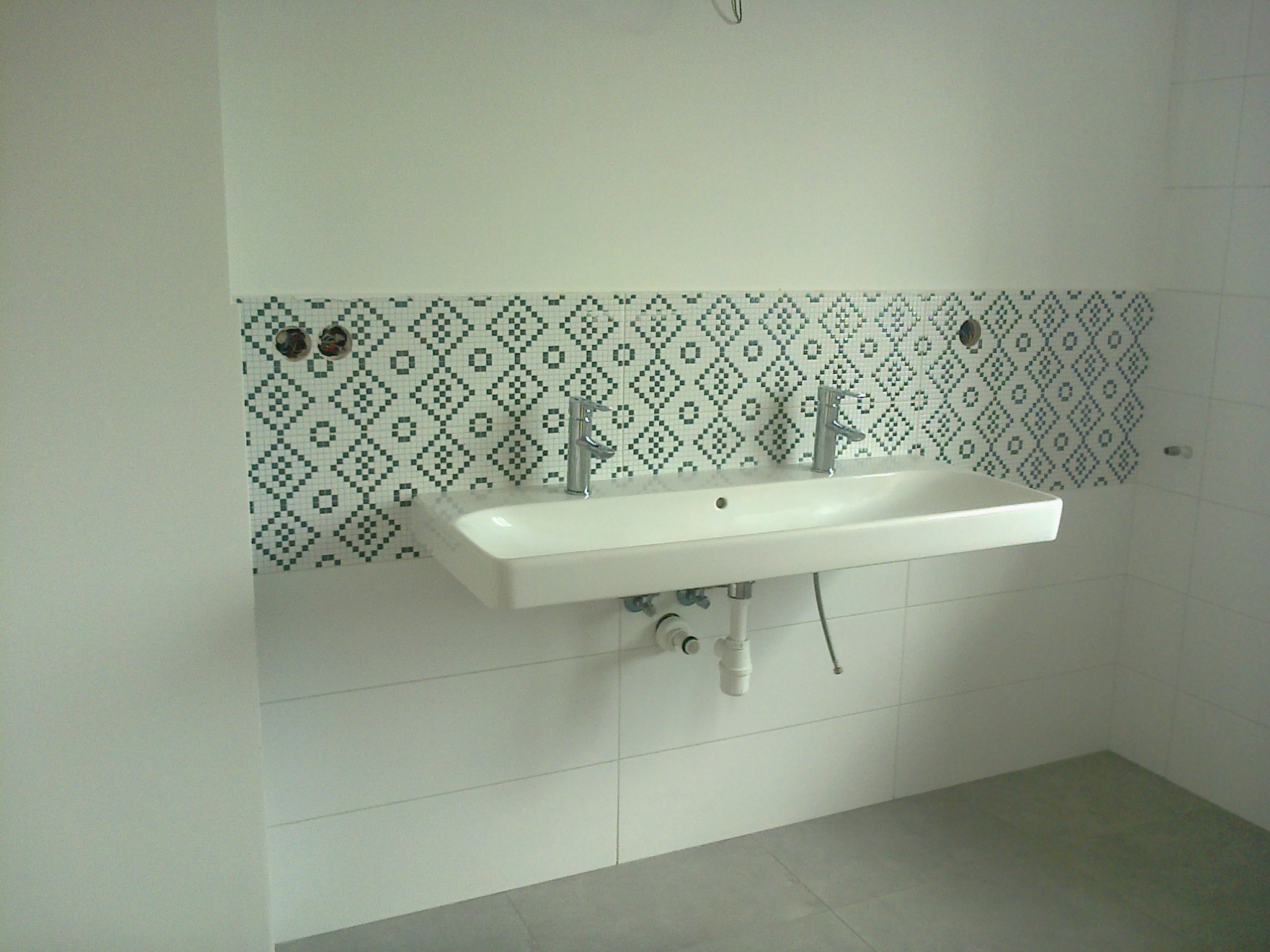 Biała umywalka z dwoma kranami zamontowana na ścianie z dekoracyjnymi mozaikami w łazience. Widoczne płytki podłogowe i rury odpływowe.