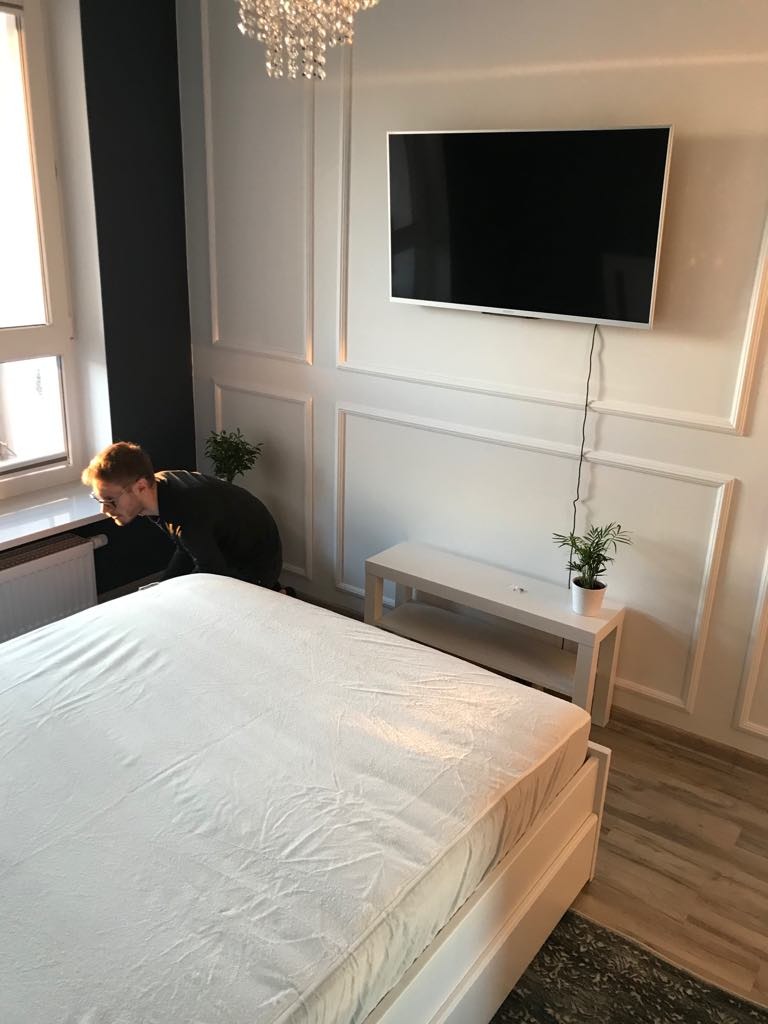 Pokój z białym łóżkiem, telewizorem na ścianie z dekoracyjnymi panelami, żyrandolem i osobą pochyloną nad oknem