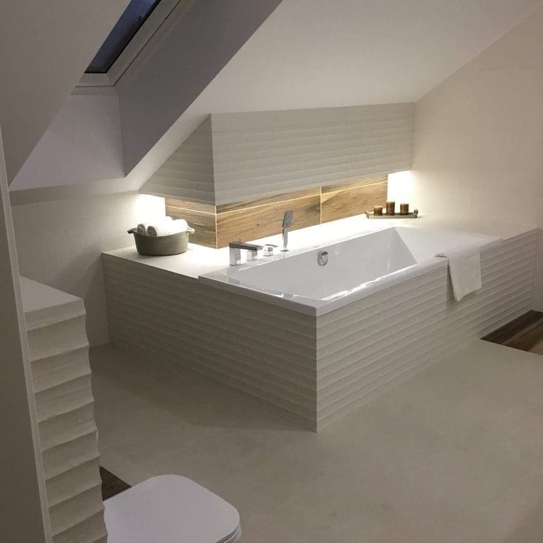 Nowoczesna łazienka na poddaszu z białą wanną obudowaną płytkami 3D, drewnopodobne płytki na ścianie, oświetlenie punktowe i koszyk z ręcznikami.