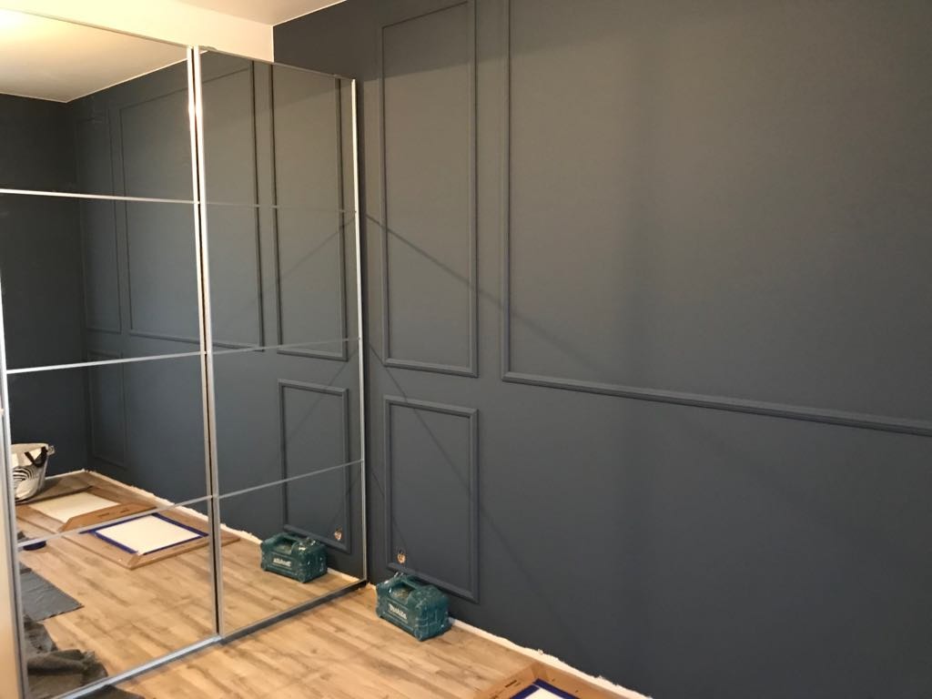 Ciemnoszara ściana z dekoracyjnymi panelami ściennymi, widoczne narzędzia i materiały przygotowane do dalszych prac wykończeniowych, odbicie w lustrzanych drzwiach szafy.