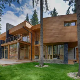 Nowoczesny dom z drewnianą elewacją i elementami z kamienia, otoczony lasem iglastym, z widocznym tarasem i balkonem ze stalową balustradą.