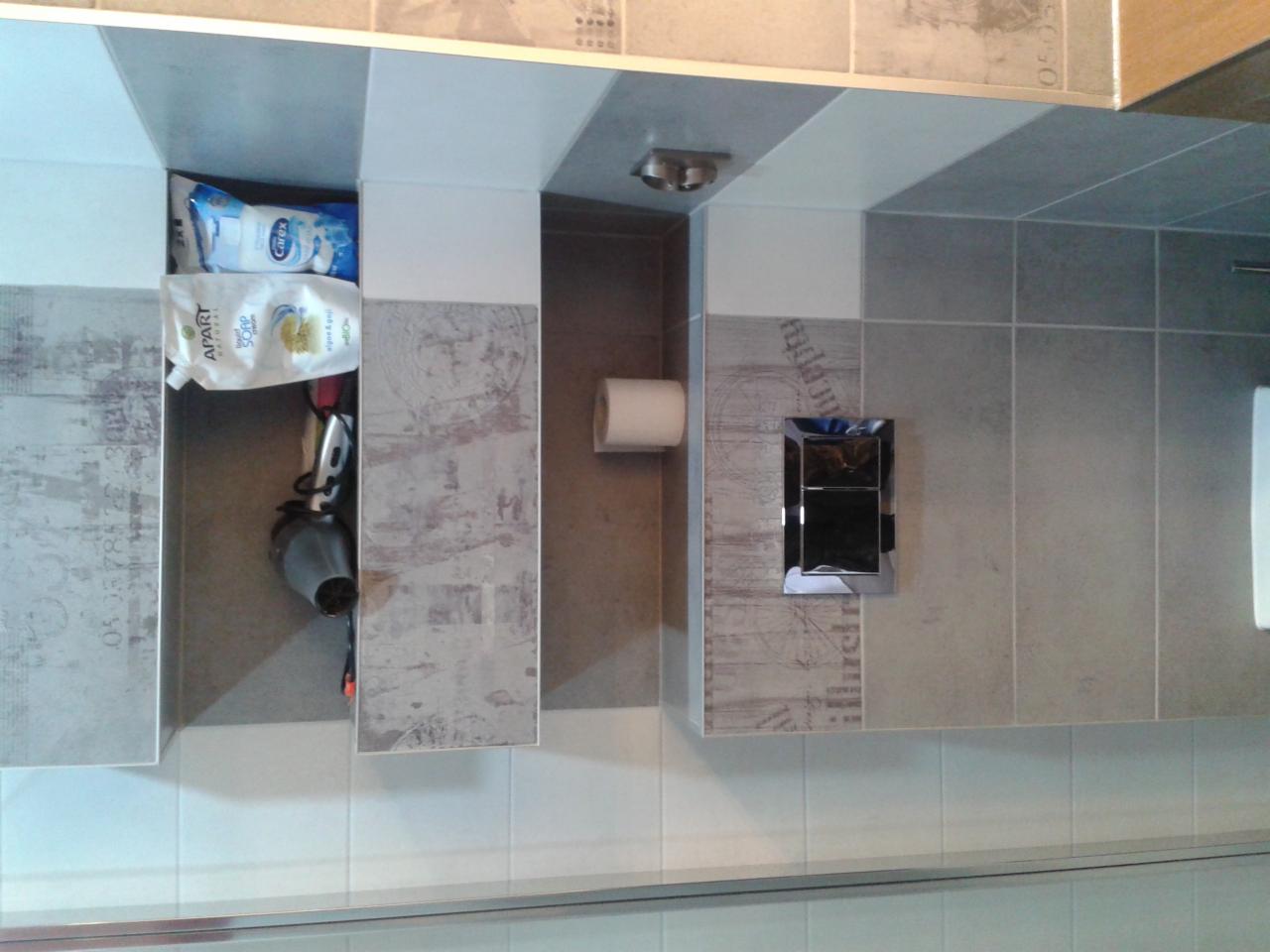 Wykończona łazienka z szarymi płytkami, wzorzystymi akcentami i wbudowanymi szafkami. Widoczny fragment toalety i akcesoria łazienkowe w szafce.