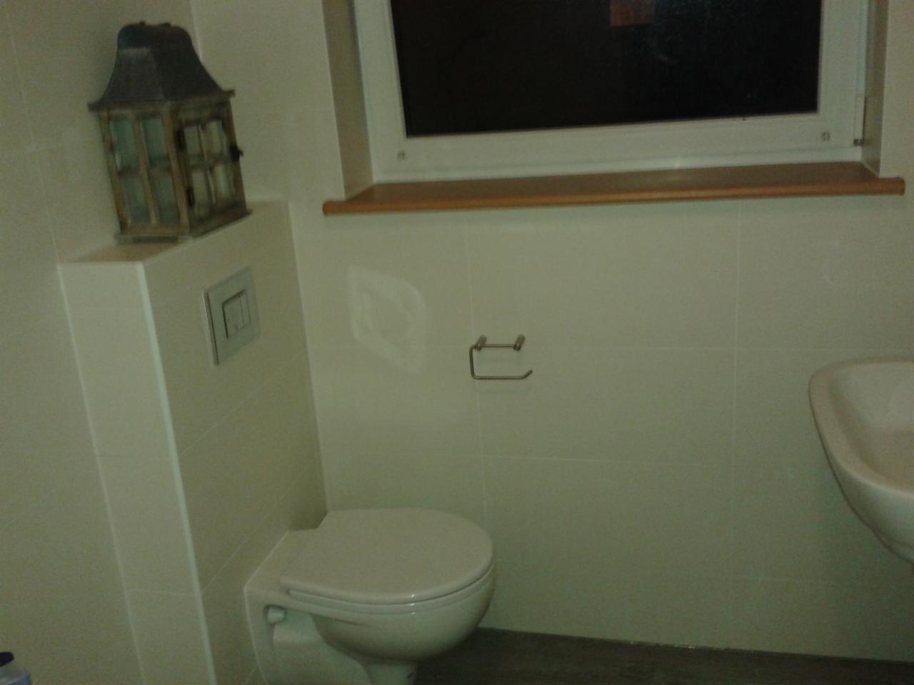 Jasna łazienka po remoncie: białe płytki, toaleta, umywalka i dekoracyjna latarenka na zabudowie geberit. Minimalistyczny design, czystość i funkcjonalność.