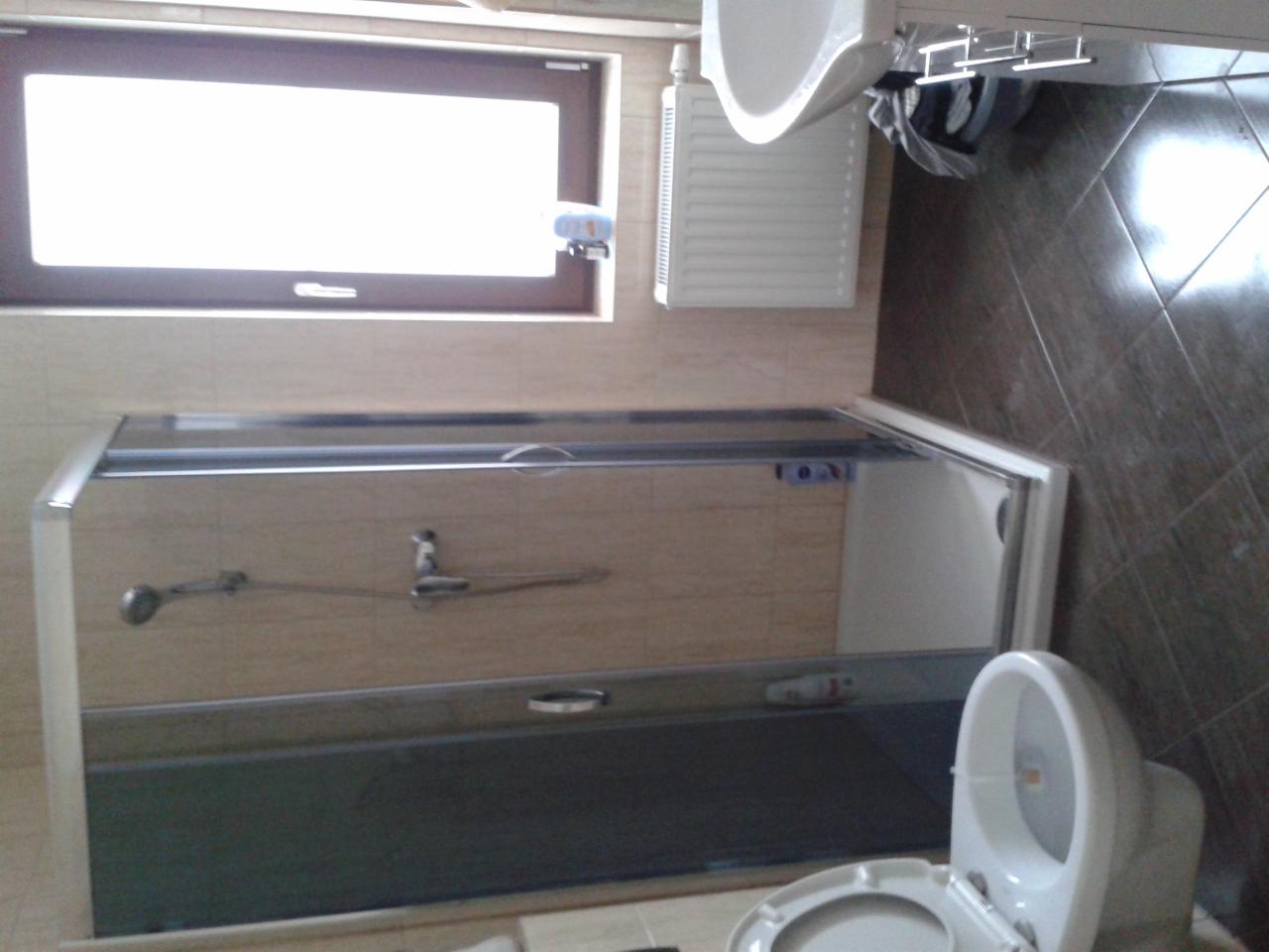 Wykończona łazienka z kabiną prysznicową, toaletą i umywalką. Widoczne płytki na ścianach i podłodze. Całość w jasnych, neutralnych barwach, z oknem w tle.