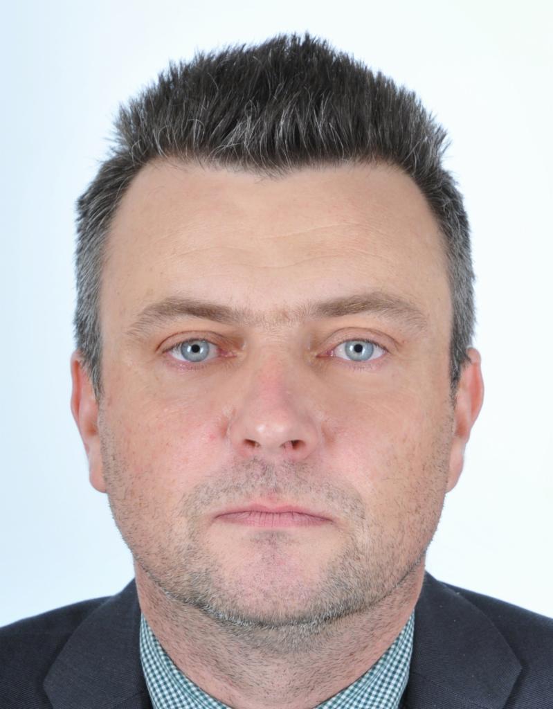 Portret mężczyzny w garniturze z jasnymi oczami na białym tle. Widoczne zmarszczki mimiczne i starannie ułożone włosy.