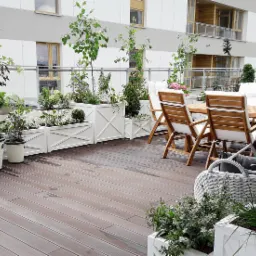 Zewnętrzny taras z drewnianą podłogą, białymi donicami z krzyżowym wzorem, różnorodną zielenią, drewnianym stołem i krzesłami oraz elementami wypoczynkowymi, na tle jasnego budynku mieszkalnego.