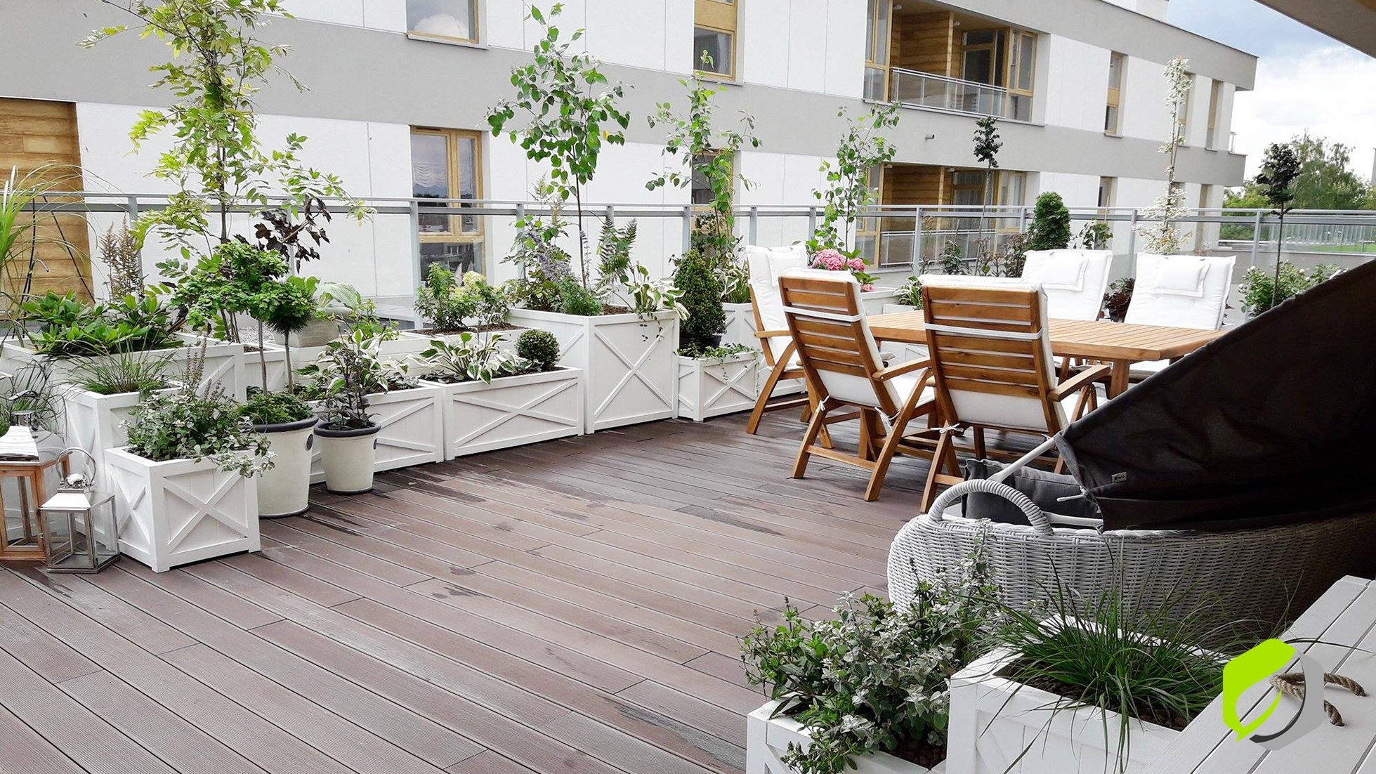 Zewnętrzny taras z drewnianą podłogą, białymi donicami z krzyżowym wzorem, różnorodną zielenią, drewnianym stołem i krzesłami oraz elementami wypoczynkowymi, na tle jasnego budynku mieszkalnego.