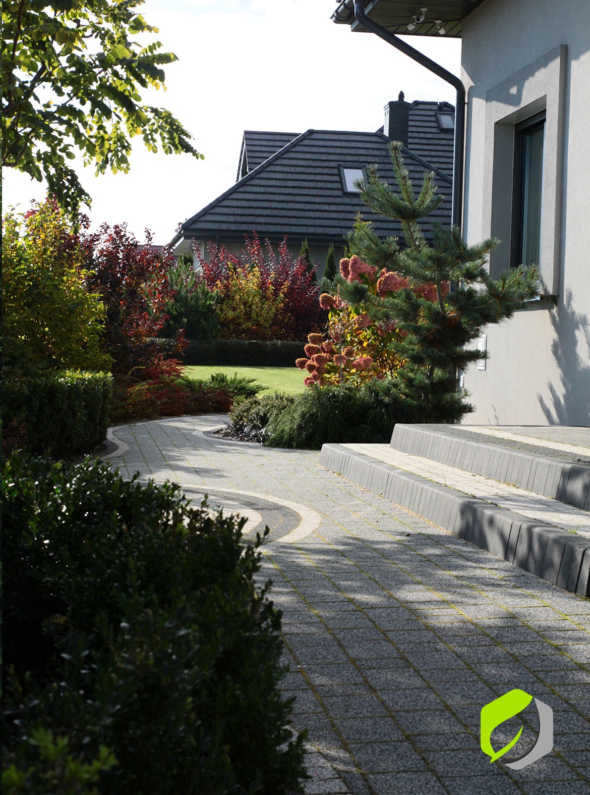 Kaskadowe schody z szarej kostki brukowej prowadzące do domu, obok ścieżka z kostki, rabaty z krzewami o jesiennych barwach i trawnik.