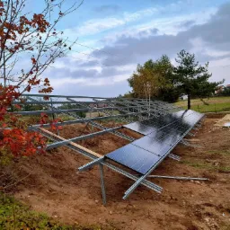 Stalowa konstrukcja wsporcza pod panele słoneczne, częściowo zamontowane panele, widok na nierówny teren, drzewa z jesiennymi liśćmi i domy w oddali pod pochmurnym niebem.