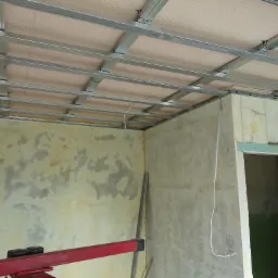 Metalowy stelaż sufitowy zamontowany w surowym pomieszczeniu, widoczne ślady po starej farbie na ścianach, przygotowanie do montażu płyt kartonowo-gipsowych.