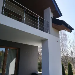 Nowoczesny dom z balkonem i elewacją w odcieniach szarości i bieli, widoczne elementy wykończeniowe, takie jak metalowa balustrada i drewniana podbitka dachu.