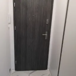 Nowe, ciemnoszare drzwi wejściowe z drewnopodobnym wzorem, widoczne od wewnątrz, z zamontowanym wizjerem i klamką, na tle białych ścian i marmurowej podłogi.